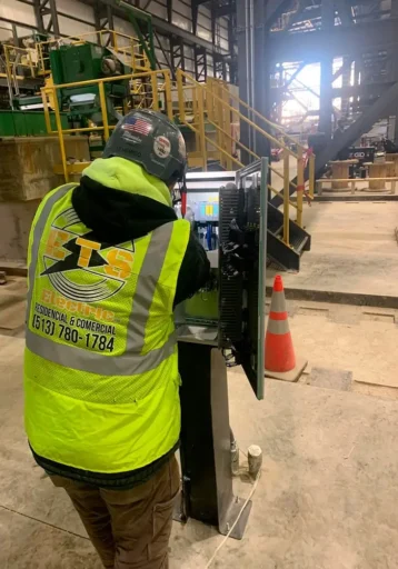 Electrician wearing a high-visibility vest and safety helmet working on an industrial control panel inside an industrial facility, surrounded by safety equipment and machinery.