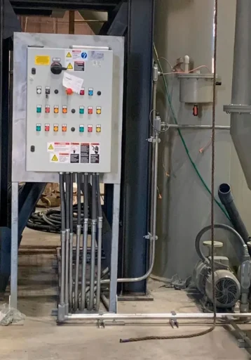 Industrial control panel mounted on a metal frame with buttons, switches, and indicator lights, connected to piping and machinery in a factory setting