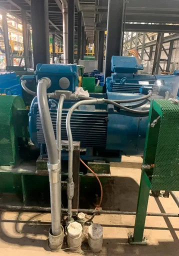 Large blue electric motors mounted on green metal structures, connected to pipes and cables in an industrial factory setting