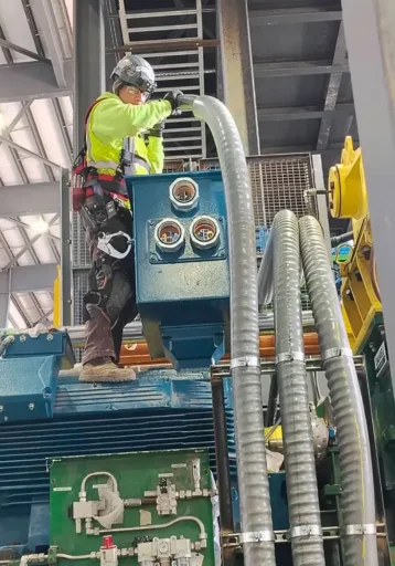 Operator in safety gear handling a flexible hose connected to a blue industrial machine in a work environment with electrical components and piping